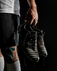 Close-up of male footballer holding cleats with sweat on hand and taped knee. Concept of post-game fatigue, recovery, emotional realism, athletic endurance, football gear.