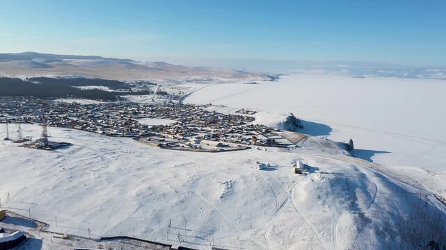 Aerial view of the snow-covered Khuzhir village, nestled beside the vast, frozen expanse of a lake under the clear blue sky, Khuzhir, Irkutsk Oblast, Russia.