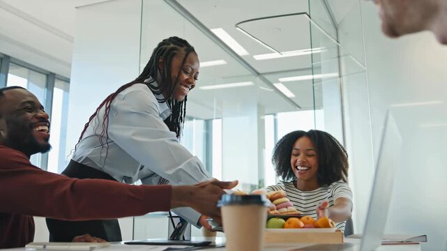 A smiling server presenting a tray of pastries and fruits in a bright, modern office setting with engaged colleagues