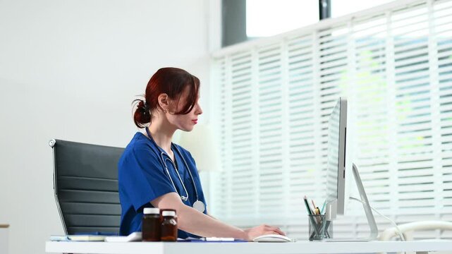 Female doctor in scrubs works at a desktop computer in bright office. Concept of modern healthcare, digital records, and admin tasks. Real people, clinical setting.