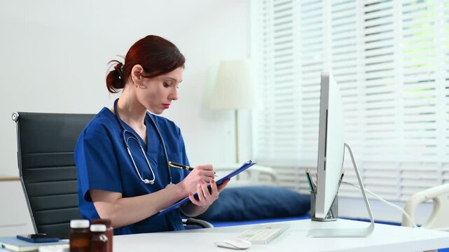 Female doctor in scrubs works at a desktop computer in bright office. Concept of modern healthcare, digital records, and admin tasks. Real people, clinical setting.