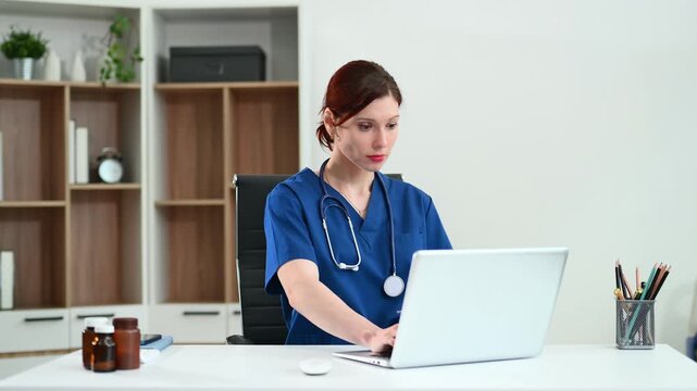 Female doctor in scrubs works at a desktop computer in bright office. Concept of modern healthcare, digital records, and admin tasks. Real people, clinical setting.