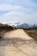 Wide gravel trail leading toward dramatic snow-covered mountains under a soft spring sky. A symbol...
