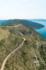 Drone photo of a winding road cutting through the sparse vegetation of Cres Island’s hilly terrain, with views over the Adriatic coastline