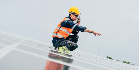 Worker inspects solar panels on rooftop during sunny day using communication device for...