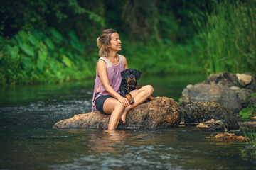 Asian woman relaxing with her Rottweiler puppy on a rock in a peaceful river. Outdoor lifestyle, pet companionship, and nature getaway concept.