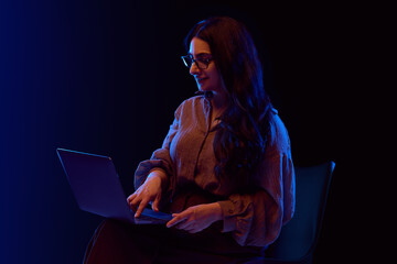 Smiling woman working on laptop in neon environment while sitting. Concept of fintech UX design,...