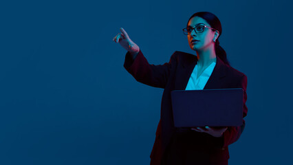 Businesswoman pointing with one hand while holding laptop under blue light. Concept of tech...