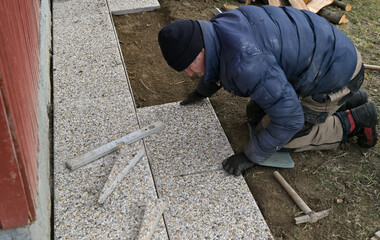 A bricklayer on his knees lays paving around a wooden house