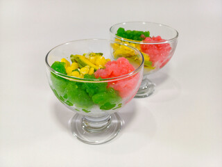 Two clear glass bowls hold vibrant portions of Es Tebak, a traditional Indonesian shaved ice dessert. Each bowl showcases layers of bright green gelatin, pale yellow corn or avocado, and pink ice