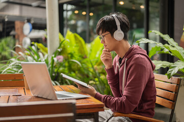 College student man wear headphone looking at notebook over laptop sitting at wooden table in a cafe