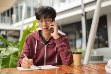 Glasses student man talking on phone writing in notebook while sitting at wooden table outside cafe.
