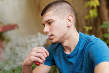 Young man vaping outdoors in a garden wearing a blue shirt during daytime