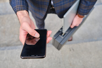 Business professional holding smartphone and laptop briefcase outdoors during work hours in an urban setting