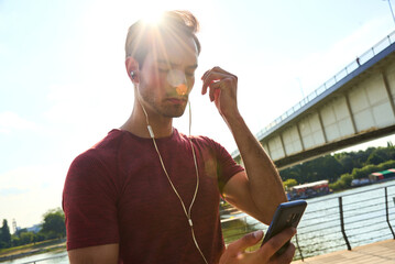 Man listens to music while using smartphone on riverbank under bright sun near bridge in summer afternoon