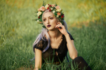 Young woman with flower crown sitting in a grassy field during sunset while wearing dark clothing and posing thoughtfully