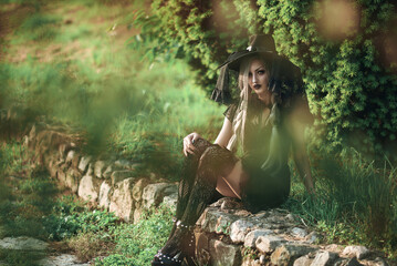 Young woman in witch costume sits on stone path surrounded by greenery during daylight