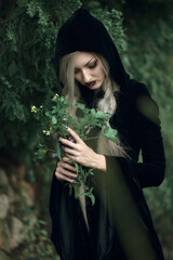 Young woman in a dark hooded outfit holding green herbs in a lush, natural setting during daytime
