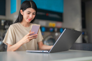Confident woman handling work and housework at once, smiling during holding or texting phone and laptop in a self-service laundry shop.