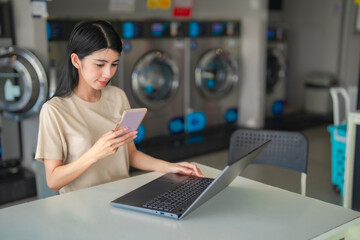 Cheerful Asian female texting on phonel while working with her laptop near a washing machine....