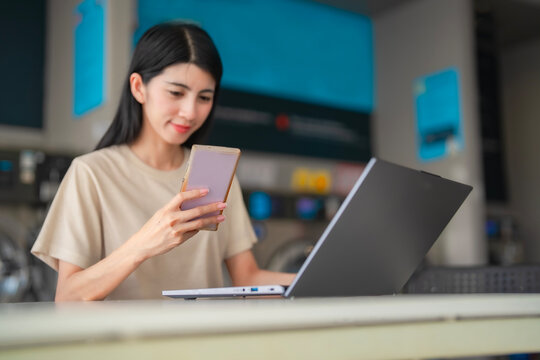 Multitasking woman doing remote work at a laundromat, managing communication and home duties efficiently. Asian freelance using smartphone and laptop at self-service laundry.