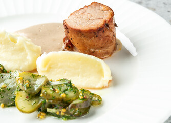 Roasted meat with creamy sauce, mashed potatoes, pickled cucumbers, and herbs served on a white...