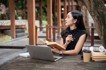 Asian woman holding pen and book looking way from laptop while sits at wooden table in outdoor cafe.