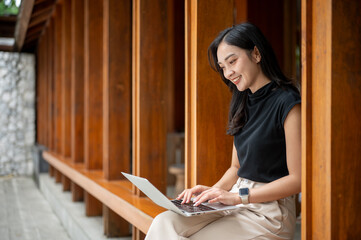 Asian woman using laptop while sitting on wooden bench in corridor or hallway of an outdoor cafe.