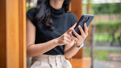 Close up woman holding and looking at phone while sit in cafe's wooden corridor beside cafe's garden