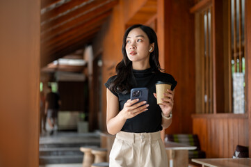 Asian woman holding phone and coffee looking around while standing or walking in cafe or restaurant.