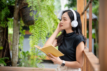 Asian woman wears headphone listening to music holding book on wood bench in cafe or garden pavilion