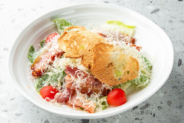 Fresh Caesar salad with crispy bacon, cherry tomatoes, Parmesan cheese, and croutons served in a white bowl on a speckled surface. Close-up food photography.