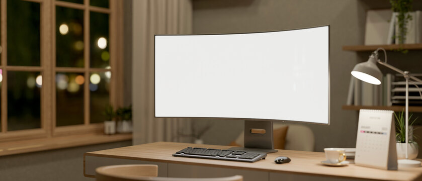 Blank curve screen computer aside coffee, calendar and lamp on wooden table in a room with bookshelf