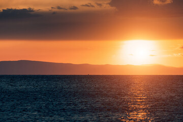 Golden sunset reflecting on calm sea with distant mountains and clouds creating a dramatic and...