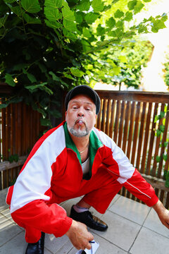 Elderly man squinting with exaggerated pout, wearing cap and bright tracksuit, humor shot.