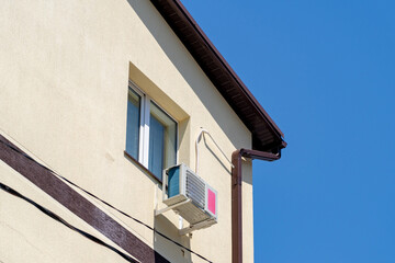 Side view of a modern building with a light beige stucco facade, featuring a window, an outdoor air conditioning unit, and brown roof gutters and downspout against a clear blue sky