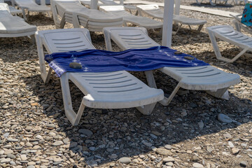 Sun-drenched pebble beach with two white plastic sun loungers, one draped with a blue towel secured...