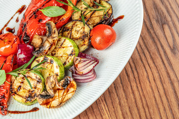 A colorful medley of grilled vegetables with fresh herbs and balsamic glaze is artfully arranged on a white ceramic plate over a wooden table. top view. copy space