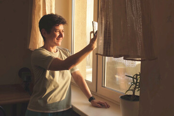 Smiling middle-aged woman raising smartphone to take a selfie at home near the window in golden...
