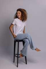 Curly Woman Sitting On Stool In White T-Shirt And Blue Jeans