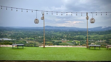 Hilltop view with green fields, hanging wooden lanterns, benches, cloudy sky, and mountain view.