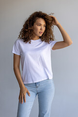 Beautiful Curly Woman Posing In White T-Shirt And Blue Jeans On Gray Background