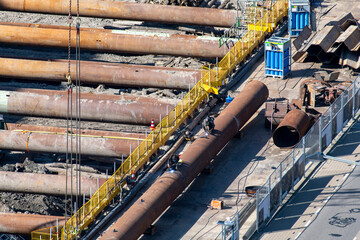 High angle view of welders working on large steel pipes for excavation pit shoring or supporting the sides of a construction site with excavation pit with sheet pile wall in background