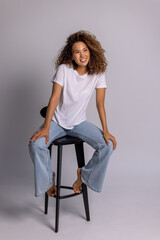 Curly Woman Sitting On Stool In White T-Shirt And Blue Jeans
