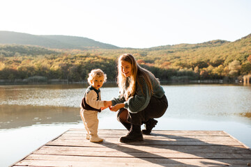 Happy woman 24-29 year old with baby girl 1-2 year old wearing knitted sweaters over nature background. Autumn season. Motherhood.