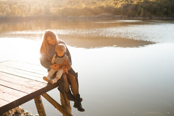 Happy woman 24-29 year old with baby girl 1-2 year old wearing knitted sweaters over lake nature background and sun light. Autumn season. Motherhood.