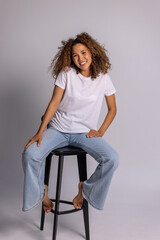 Curly Woman Sitting On Stool In White T-Shirt And Blue Jeans