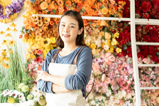 Asian female florist in apron smiling while designing a beautiful floral arrangement with her colleague in a flower shop. Concept of teamwork, creativity, floral business, and joyful workplace. - Powered by Adobe