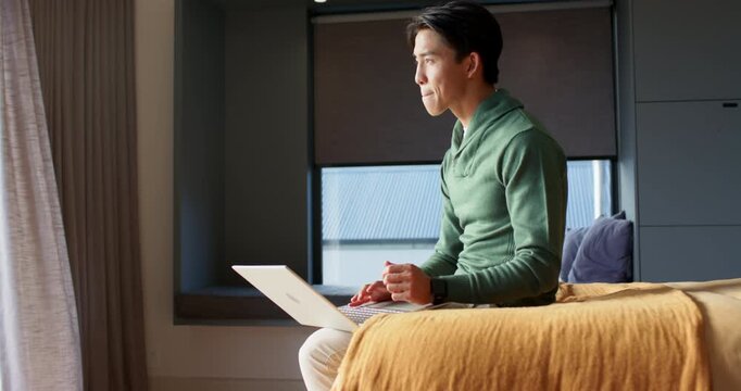 Asian man sitting on bed placing silver laptop on lap, typing then closing, celebrating success