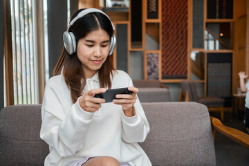 A young woman wearing headphones, enjoying music or gaming on her smartphone while seated in a stylish cafe.